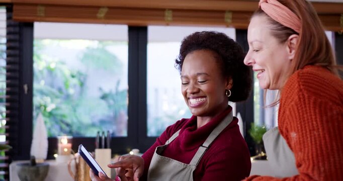 Diverse female friends wearing aprons in kitchen, scrolling tablet showing recipe win, high-fiving