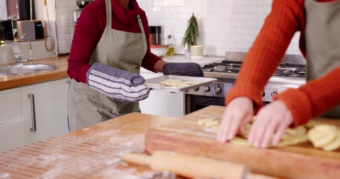 Female friends baking, oven timer dinging, woman in maroon with mitts presenting cookies to partner