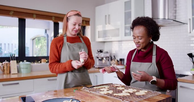 Diverse female friends squeezing bags, decorating cookies on rack at kitchen island wearing aprons