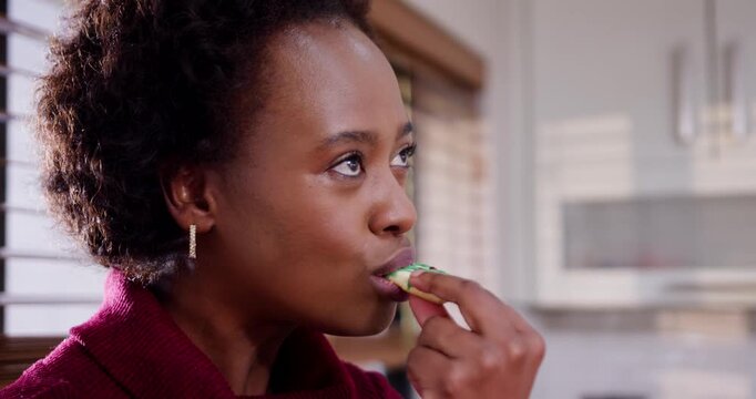 African woman wearing burgundy knit raising green-iced cookie to lips, tasting tea in kitchen