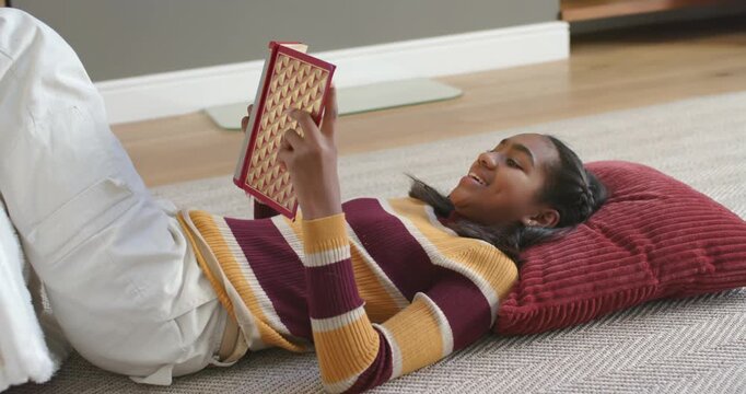Female teenager lying at home on rug in striped sweater, holding red e-reader, reading and laughing