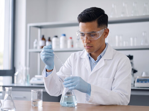young hispanic male student wearing lab coat and goggles using pipette with conical flask for chemistry analysis in university laboratory