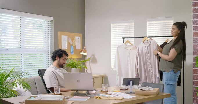 African American partners, woman presenting shirts for feedback while man reviewing laptop at desk