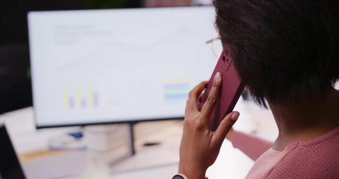On call African American woman at desk holding maroon cell viewing monitor reading data, copy space
