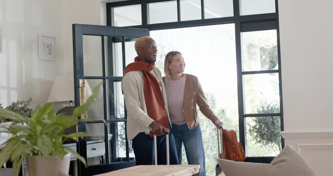 Diverse couple arriving through open glass door, carrying wheeled suitcases and orange backpack