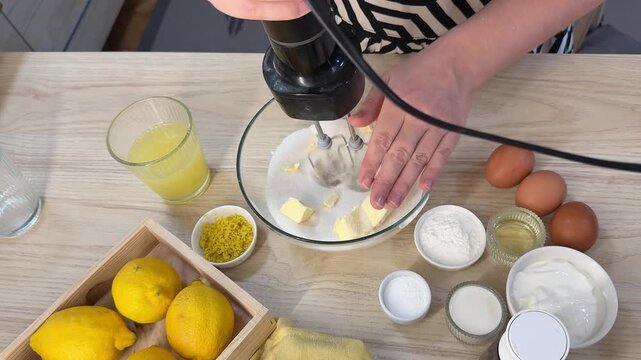 A person uses an electric hand mixer to blend butter and sugar in a glass bowl, surrounded by lemons, eggs, flour, and cream on a wooden table