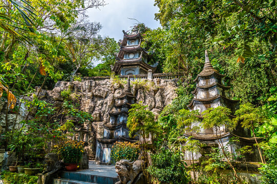 A view towards a tiered pagoda nestled against a rocky cliff face surrounded by lush greenery at the Marble Mountains in Da Nang, Vietnam on a bright sunny day