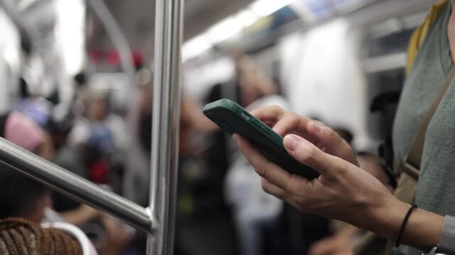 Close-up of a commuter's hands texting on a smartphone while traveling on a crowded new york city subway, with passengers in the background holding onto the handrail