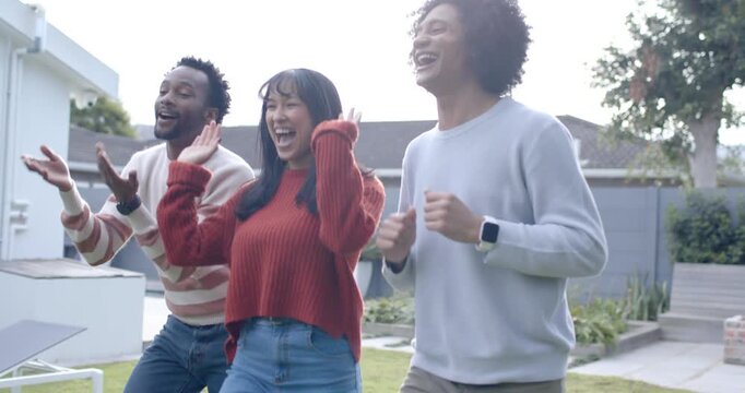 Diverse friends stepping and laughing on suburban yard by pool, woman in red sweater raising hands