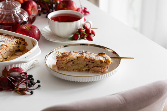Appealing slice of apple pie with hot tea served on a white table surrounded by fresh apples and autumn leaves