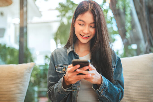 Smiling young woman using smartphone while sitting comfortably on sofa in bright modern space, enjoying mobile technology, online connection.