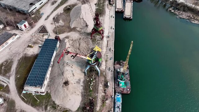 Rail-mounted portal cranes and grab bucket at an aggregate quay in steep overhead drone view. Bulk handling, river-port layout and construction aggregate logistics