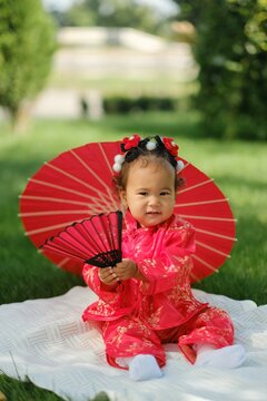 Asian baby girl in traditional red dress holding fan with red umbrella outdoors on grass