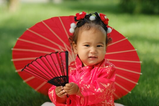 Asian baby girl in traditional red dress holding fan with red umbrella outdoors on grass