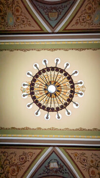 Low angle bottom-up view of an elegant round vintage chandelier with glowing lights installed on a decorated ceiling with classic frescoes and gold ornamentation.