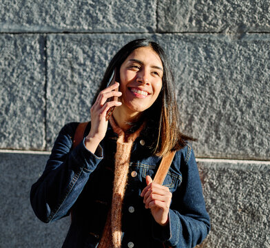 Young woman stands against a stone wall, smiling as she talks on her phone. Casual denim jacket and cozy sweater suggest a relaxed, urban lifestyle moment. Natural sunlight highlights her face and hai