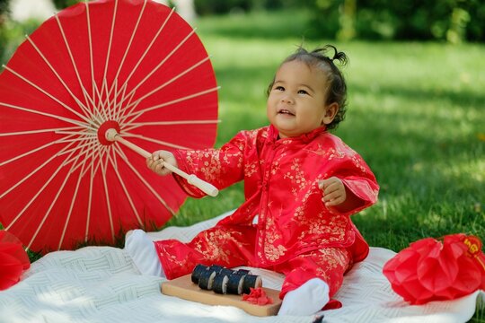 Asian baby in traditional red costume with umbrella, sushi rolls and lantern on picnic blanket