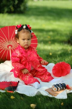 Asian baby in traditional red costume with umbrella, sushi rolls and lantern on picnic blanket