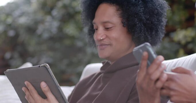 Diverse friends laughing, adult male of African descent scrolling tablet on outdoor sofa, tapping