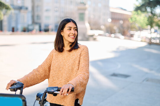 A young woman smiles as she rides a scooter along a tree-lined city street, wearing a cozy orange sweater. The image conveys casual urban lifestyle, contentment and autumn warmth, suitable for travel,