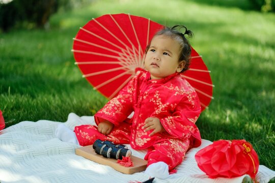 Asian baby in traditional red costume with umbrella, sushi rolls and lantern on picnic blanket
