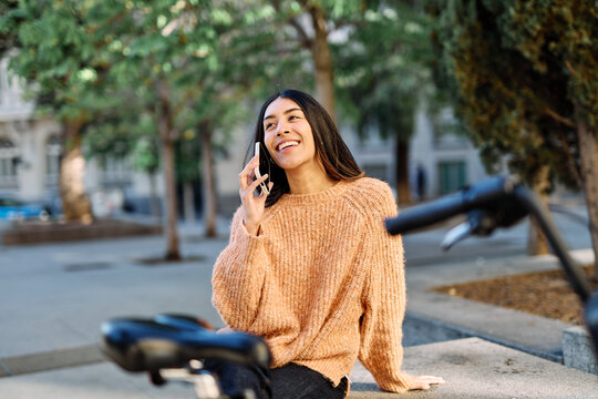 A young woman sits by a parked bicycle in an urban park, holding a smartphone and gazing thoughtfully into the distance. She wears a cozy sweater and jeans, suggesting a relaxed autumn outing. The can