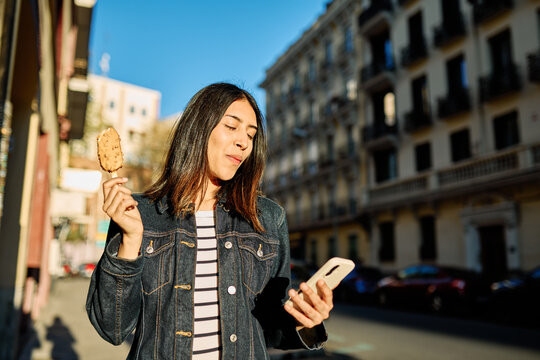 Portrait of a smiling young woman girl eating an ice cream and looking at her phone walking in the city, tourists visiting destination, summer trip exploring