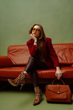 Fashionable woman wearing elegant burgundy red blazer, leather leggins, leopard print ankle boots, eyeglasses, holding brown bag, sitting on sofa, posing on green background. Full-length portrait