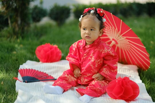 Asian baby in traditional red suit sitting on picnic blanket with fan, umbrella and paper lanterns
