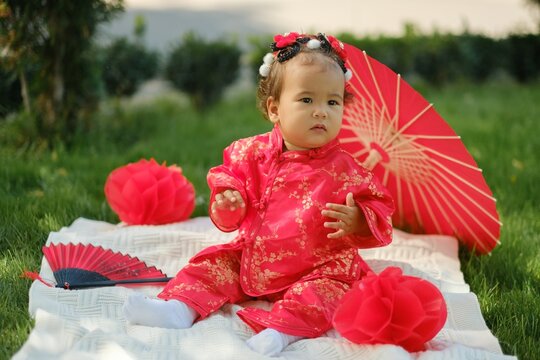 Asian baby in traditional red suit sitting on picnic blanket with fan, umbrella and paper lanterns