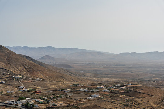A scattered village of white and terracotta-roofed houses nestles at the base of a dark volcanic mountain, overlooking a hazy arid valley, Fuerteventura, Canary Islands, Spain.