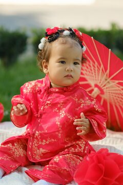Asian baby in traditional red suit sitting on picnic blanket with fan, umbrella and paper lanterns