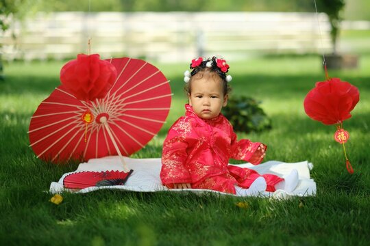 Asian baby in traditional red suit sitting on picnic blanket with fan, umbrella and paper lanterns