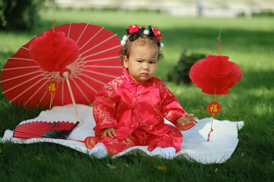 Asian baby in traditional red suit sitting on picnic blanket with fan, umbrella and paper lanterns