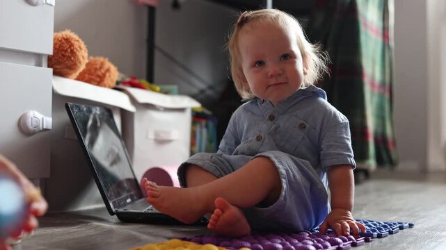 A curious toddler sitting on the floor in a bright room with a laptop and colorful toys nearby