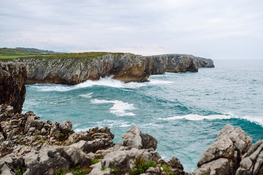 Waves crash against rocky cliffs at the water's edge. Painting depicts ocean water striking rocks and sending up spray, set against a gray sky. Concept of wild nature; a peaceful landscape.