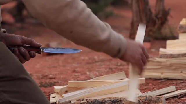 Outdoor carpentry moment hands shape wooden stake with knife beside lumber stack under warm daylight in rustic workshop setting showing texture of wood and careful shaving