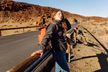 Female traveler with backpack leans against roadside barrier in remote, open landscape. Woman is resting at sunset in national park near volcano. Adventure and wilderness themes. © maxbelchenko