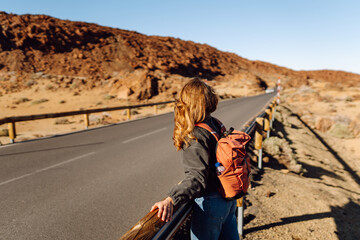 Female traveler with backpack leans against roadside barrier in remote, open landscape. Woman is resting at sunset in national park near volcano. Adventure and wilderness themes. © maxbelchenko