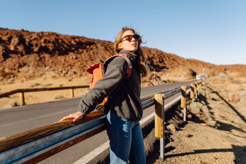 Female traveler with backpack leans against roadside barrier in remote, open landscape. Woman is resting at sunset in national park near volcano. Adventure and wilderness themes. © maxbelchenko