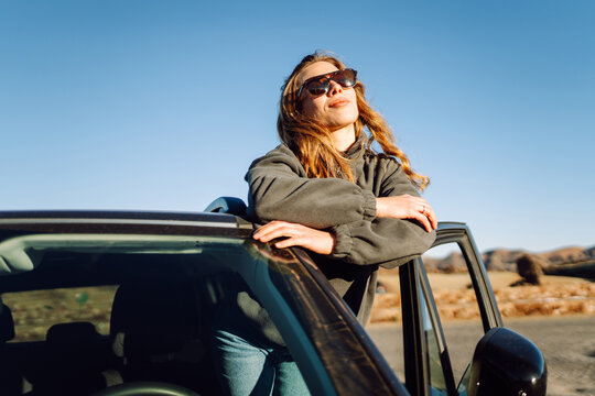 Woman wearing sunglasses enjoys desert scenery near volcano at sunset during a road trip. Female traveler with backpack stands near her car in a national park. Nature and adventure concept.