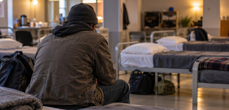 Homeless man sitting on bed in homeless shelter. Male with backpack resting in dormitory with rows of beds. Social support, poverty and charity concept.
