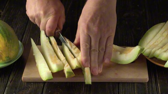 Hands slicing green melon into thin pieces on a wooden cutting board, with a whole melon and a plate of cut pieces visible in the background, showcasing the preparation process in a kitchen setting
