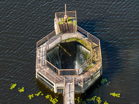 Aerial view of a hexagonal water intake structure with a metal railing and cascading water in a lake surrounded by lily pads in Plunge, Telsiai County, Lithuania.