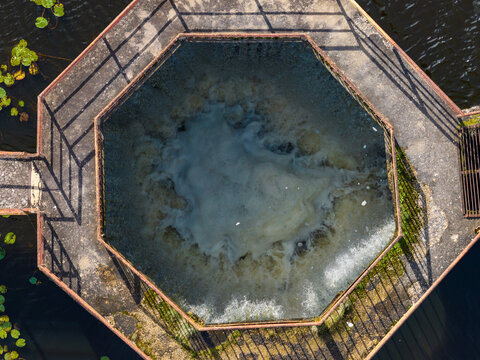 Aerial view of an octagonal water spillway structure with churning water, surrounded by a concrete walkway and lily pads, Plunge, Telsiai County, Lithuania.