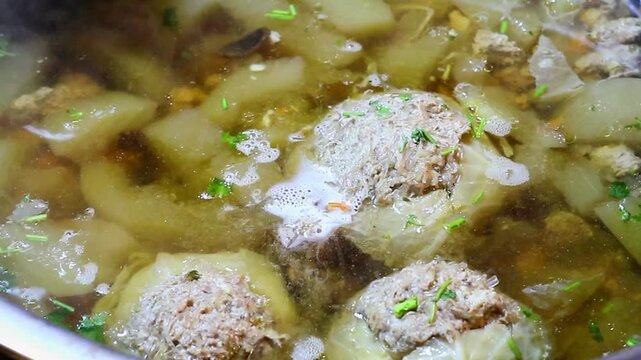 Close up of stuffed cabbage rolls and winter melon simmering gently in a metal pot, with light bubbles and steam rising during the cooking process.