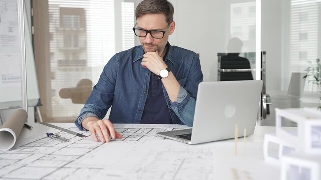 Male architect planning new construction project, creating building blueprint plans, calculating dimensions, and working on a laptop in a modern design office
