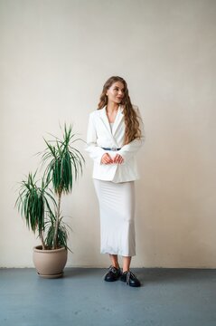 Young woman in white blazer and fitted skirt standing beside tall potted dracaena plant against neutral studio wall