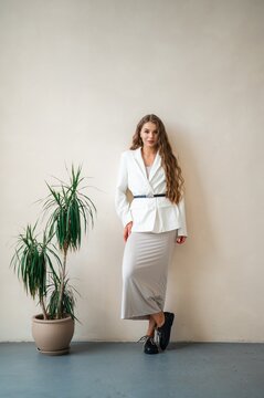 Young woman in white blazer and long grey skirt posing beside potted dracaena on neutral studio wall