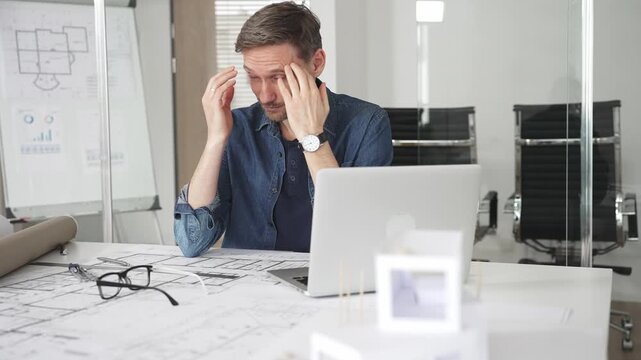 Stressed architect feeling overwhelmed and frustrated, suffering from burnout while working on blueprints for a tight project deadline in a modern office setup